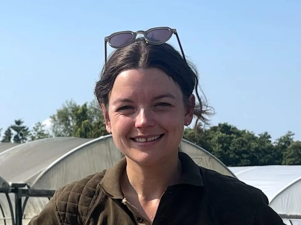 Hetty Henley smiling, sunglasses pushed up, by a nursery polytunnel